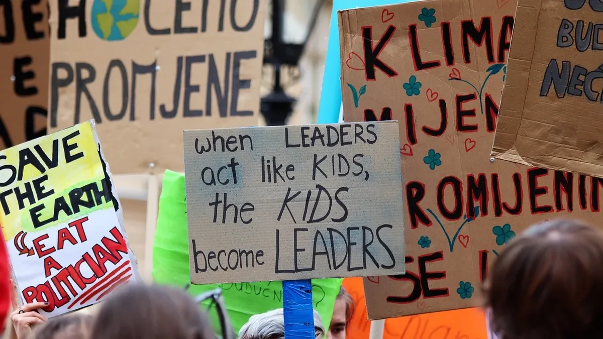 A group of people holding signs with messages about climate change and leadership, set against a backdrop of other signs and a street lamp.